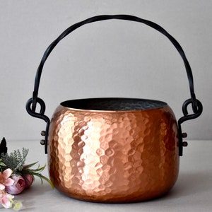 May include: A hammered copper pot with a black wrought iron handle. The pot has a rounded shape and a textured surface.