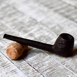 Pode incluir: Um cachimbo de tabaco castanho escuro com uma tigela texturizada e um longo caule repousa sobre uma rolha. O cachimbo está sobre uma superfície de madeira clara e envelhecida, criando uma estética rústica.