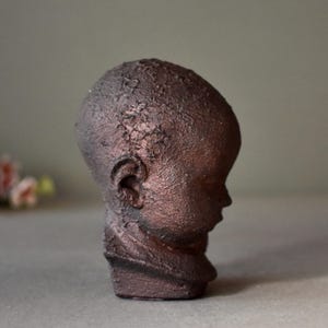 May include: A small, brown ceramic sculpture of a child's head. The sculpture has a textured surface and is in a profile view.