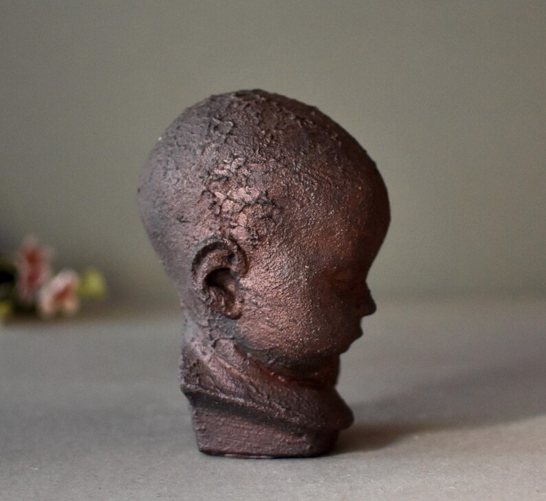 May include: A small, brown ceramic sculpture of a child's head. The sculpture has a textured surface and is in a profile view.