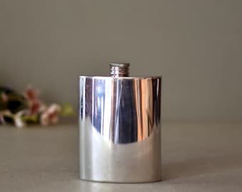 Puede incluir: Una petaca de plata con forma rectangular redondeada y un diseño simple y liso.