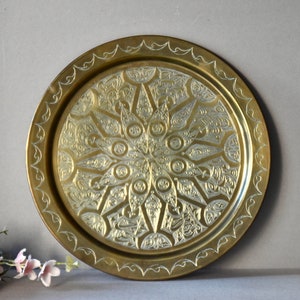May include: A round brass serving tray with an intricate, embossed, silver-toned design. The tray has a raised rim with a decorative border.