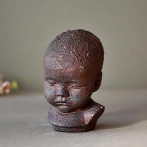 May include: A small ceramic bust of a child with a dark brown glaze. The bust is sculpted with a realistic expression and features a textured surface.