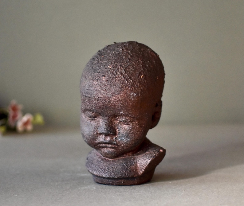 May include: A small ceramic bust of a child with a dark brown glaze. The bust is sculpted with a realistic expression and features a textured surface.