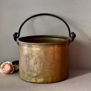 May include: A vintage brass pot with a black metal handle. The pot is round and has a worn, aged look.
