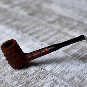 Pode incluir: Um cachimbo de madeira marrom com um caule preto. O cachimbo tem um design entalhado e está apoiado em uma superfície de madeira branca.