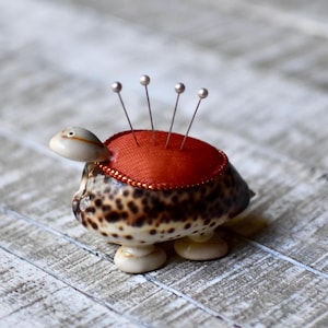 Puede incluir: Un original cojín para alfileres con forma de tortuga. El caparazón es de color marrón y crema moteado. La parte posterior de la tortuga es un cojín rojo con alfileres plateados. La tortuga tiene cabeza, patas y cola color crema.