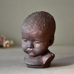May include: A small ceramic sculpture of a child's head with a textured, brown finish. The sculpture has a realistic expression with closed eyes and a slightly downturned mouth.