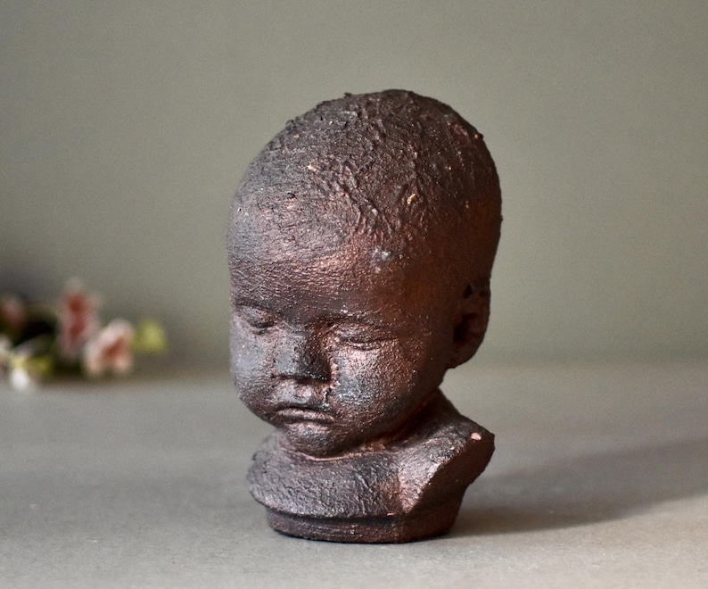 May include: A small ceramic sculpture of a child's head with a textured, brown finish. The sculpture has a realistic expression with closed eyes and a slightly downturned mouth.