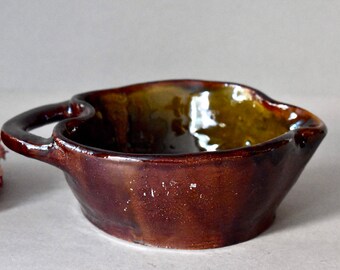 May include: A small, brown ceramic bowl with a green glaze on the inside. The bowl has two handles and a slightly irregular shape.