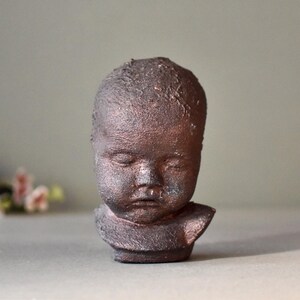 May include: A small ceramic sculpture of a child's head with a textured surface. The sculpture is painted in a dark brown color.