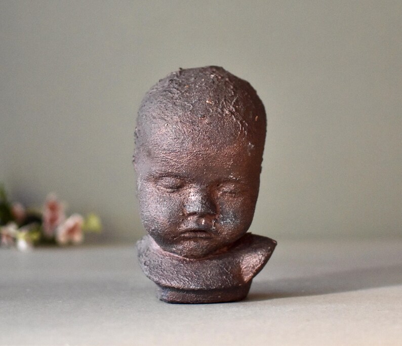 May include: A small ceramic sculpture of a child's head with a textured surface. The sculpture is painted in a dark brown color.