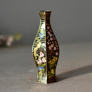 May include: A small, decorative vase with a black background and gold trim. The vase is adorned with floral patterns in pink, blue, and yellow. The vase has a unique shape and is likely made of metal.