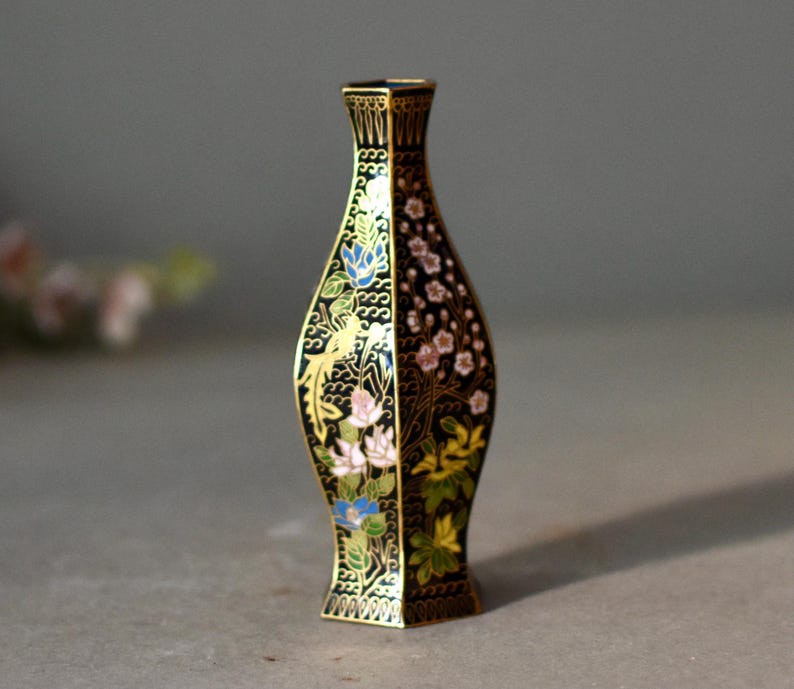 May include: A small, decorative vase with a black background and gold trim. The vase is adorned with floral patterns in pink, blue, and yellow. The vase has a unique shape and is likely made of metal.