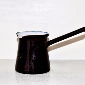 Puede incluir: Una cafetera de esmalte negro con interior blanco y diseño floral. La cafetera tiene un asa larga y un pico.