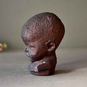 May include: A ceramic sculpture of a child's head with a textured, dark brown finish. The sculpture is in a profile view, with the child's head tilted slightly down.