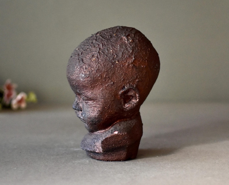 May include: A ceramic sculpture of a child's head with a textured, dark brown finish. The sculpture is in a profile view, with the child's head tilted slightly down.