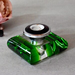 May include: A green glass bottle with a clear glass stopper and a crystal top. The bottle has a square base and a rounded top.