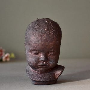 May include: A small ceramic sculpture of a baby's head with closed eyes. The sculpture is made of dark brown clay and has a textured surface.