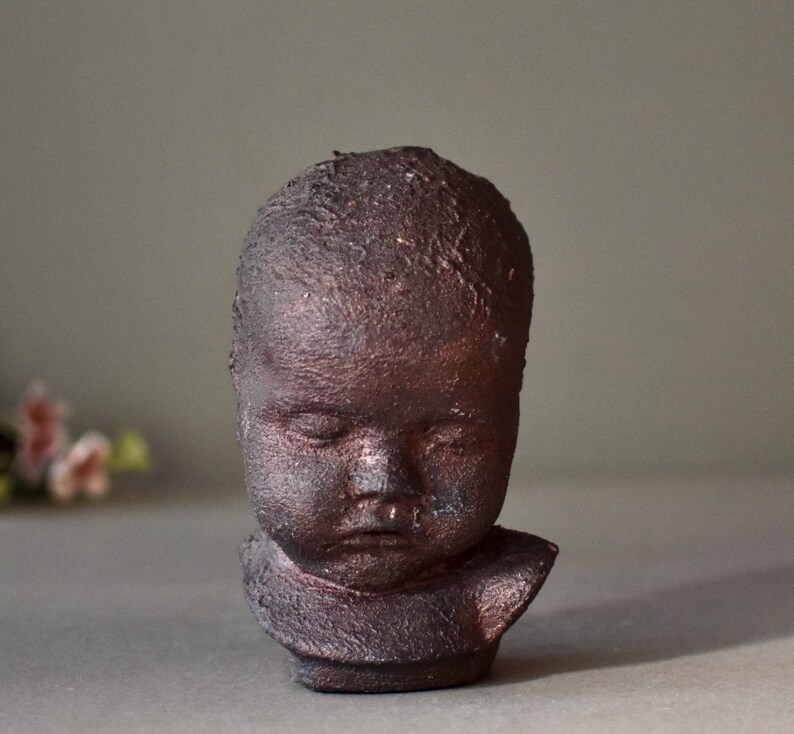 May include: A small ceramic sculpture of a baby's head with closed eyes. The sculpture is made of dark brown clay and has a textured surface.
