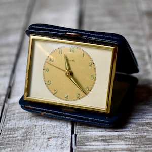Può includere: Una sveglia da viaggio rettangolare con quadrante dorato e lancette metalliche, custodita in una custodia pieghevole blu scuro simile alla pelle. L'orologio presenta un quadrante circolare con numeri arabi e una cornice color oro.