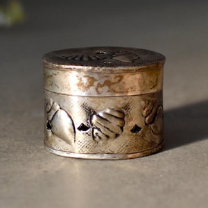 以下が含まれることがあります： 蓋付きの小さな円筒形の銀色の箱。箱はエンボス加工された花と葉のデザインで装飾されています。金属は変色した外観をしており、古さを感じさせます。箱は小さなアイテムを保管するために使用される可能性があります。