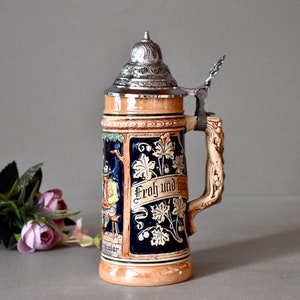 May include: A ceramic beer stein with a pewter lid and handle. The stein is decorated with a blue and white floral design and the text "Froh und frei".