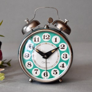 Può includere: Una sveglia vintage verde con campane argentate e lancette nere. Il quadrante dell'orologio ha numeri bianchi e uno sfondo bianco con un bordo dorato. L'orologio indica "Сделано в СССР" che si traduce in "Made in URSS".