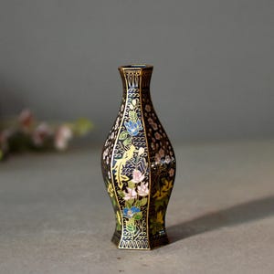 May include: A black decorative vase with a floral and bird design. The vase has a gold rim and base, with colorful flowers and birds. The vase is approximately 6 inches tall.