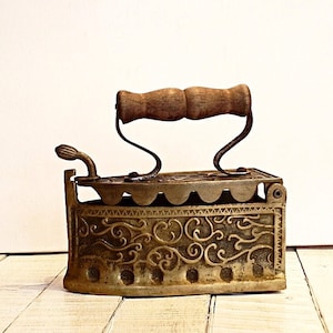 May include: Antique metal iron with a wooden handle. The iron has a decorative, ornate design on the front and sides. The handle is made of wood and is attached to the iron with curved metal supports. The iron is a vintage item, likely used for pressing clothes.