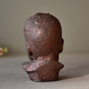 May include: A terracotta sculpture of a person's head and shoulders. The sculpture is a dark brown color and has a rough, textured surface.