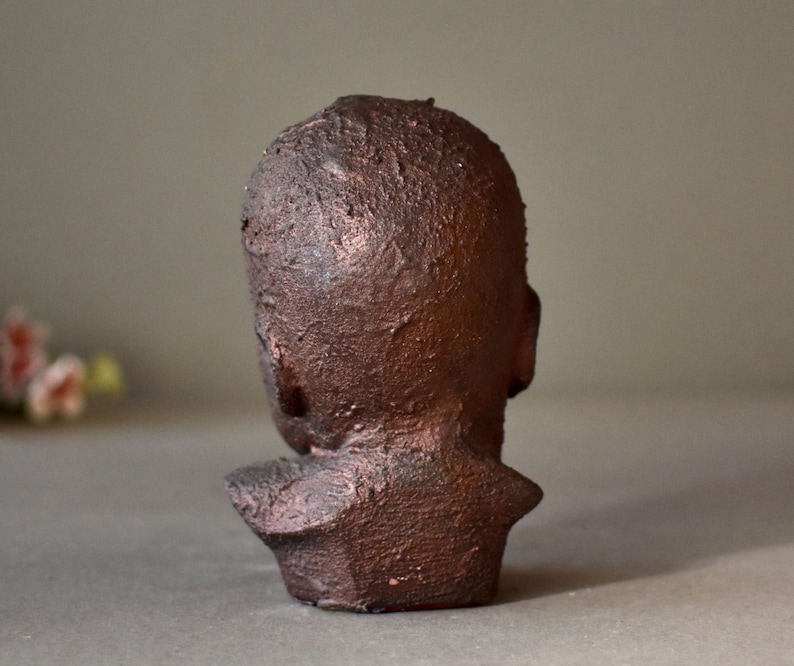 May include: A terracotta sculpture of a person's head and shoulders. The sculpture is a dark brown color and has a rough, textured surface.