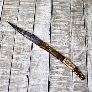 May include: An antique knife with a long, pointed blade and a decorative handle. The handle features brass accents, a cream-colored section with an etched floral design, and a series of brass circles at the end. The blade shows signs of age and wear.