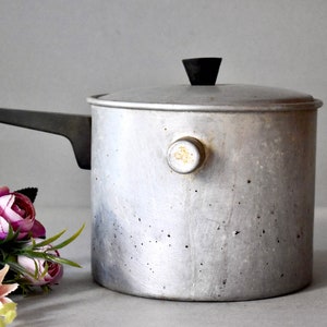 May include: A silver aluminum saucepan with a black lid and handle. The saucepan has a small knob on the side and shows signs of wear.