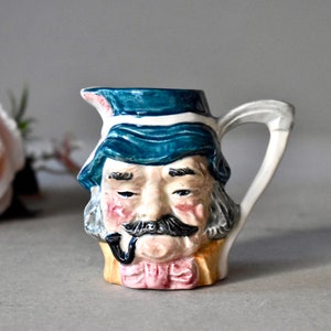 Può includere: Un piccolo brocca di ceramica a forma di testa d'uomo. La brocca ha un cappello blu, un baffo bianco e una pipa. La brocca è dipinta in tonalità di blu, bianco e rosa.