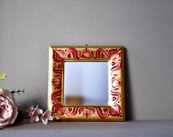Puede incluir: Un pequeño marco de imagen pintado en oro con un diseño floral rojo y dorado. El marco tiene un espejo en el interior.