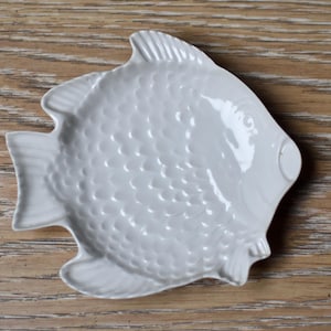 Könnte beinhalten: Eine weiße Keramikschale in Fischform mit glänzender Oberfläche und geprägtem Schuppenmuster auf dem Körper. Das Design umfasst ausgeprägte Flossen, einen Schwanz und ein kreisförmiges Augendetail, was ein funktionales Objekt mit glatter Form ergibt.