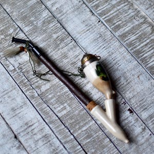 May include: A vintage porcelain pipe with a brown wooden stem and a white and green floral design. The pipe has a small metal cap on the top.