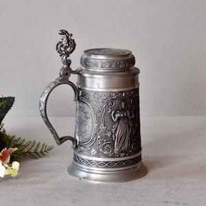 May include: A silver pewter beer stein with an ornate lid and handle. The stein features an embossed design of a woman holding a pitcher and grapes. The stein is decorated with floral motifs and a band of rope.