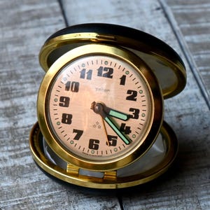 Könnte beinhalten: Eine Vintage-Reiseuhr mit einem goldfarbenen Gehäuse und einem hellbraunen Zifferblatt. Die Uhr hat schwarze Ziffern, grüne Zeiger und das Wort "peter" auf dem Zifferblatt. Die Uhr ist geöffnet und zeigt das Zifferblatt und die Glasabdeckung.