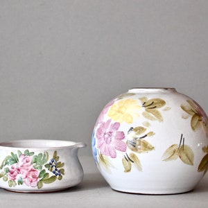 May include: Two white ceramic vases with floral designs. The larger vase is round and has a pink, yellow, and green floral pattern. The smaller vase is a bowl shape with a pink and green floral pattern.