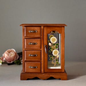 May include: A wooden jewelry box with four drawers and a glass door. The door features a floral design with yellow flowers and green leaves.