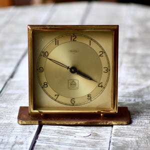 Op de afbeelding: Een vintage vierkante klok in messingkleur met een gouden wijzerplaat en zwarte wijzers. De klok heeft een rechthoekige basis en een gouden frame. De wijzerplaat heeft zwarte cijfers en de merknaam "Hovet".