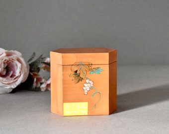 May include: A hexagonal wooden box with a light brown finish and a painted design of a vine with green leaves and pink grapes. The box has a yellow label with the text "Japanese Tea" in black.