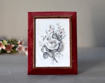 May include: A red velvet picture frame with a gold trim, holding a vintage floral print of a white rose and other flowers.