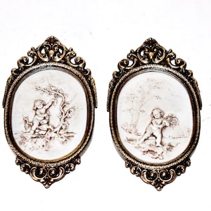 Può includere: Due placche da parete ovali con cornici dorate ornate. Ogni placca presenta uno sfondo bianco con una scultura in rilievo marrone di un cherubino in un ambiente boschivo.