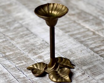 Puede incluir: Un candelero de latón con diseño floral. El candelero tiene una copa redonda para la vela, un tallo largo y tres pies en forma de hojas. El latón tiene un acabado cálido y antiguo, y el diseño es elegante y decorativo.