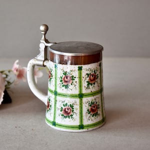 May include: A white ceramic beer stein with a green and blue floral pattern. The stein has a hinged lid with a silver metal handle.