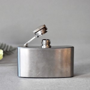 May include: A silver flask with a hinged cap. The cap is open and the flask is sitting on a gray surface.