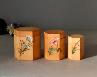 May include: Three wooden hexagonal boxes of varying sizes, each with a floral design. The boxes are light brown with hand-painted flowers in blue, pink, and red. The boxes are likely for storage or decoration.